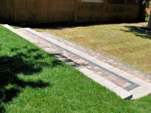 Backyard stone walkway with multicolored pavers separates green grass from a dry patch, leading to a wooden privacy fence.
