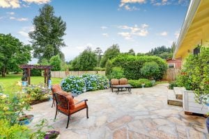Backyard stone patio with cushioned chairs, blooming hydrangeas, lush hedges, and wooden arbor under a partly cloudy sky.