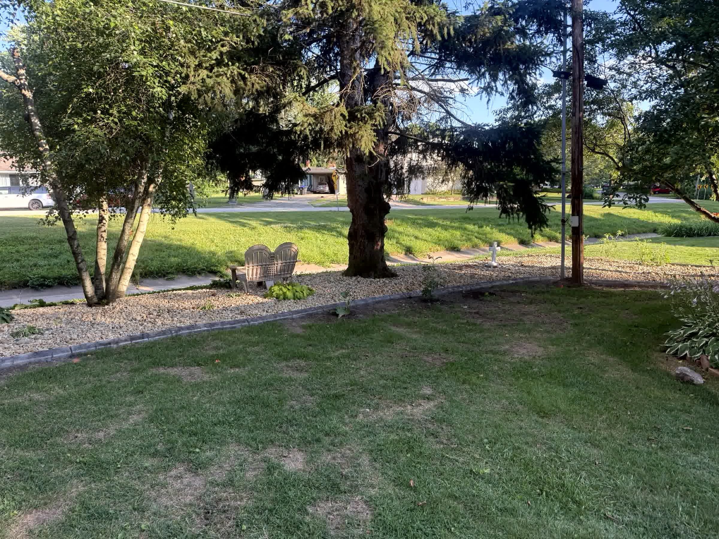 Shaded front yard with mature trees, gravel border, small garden plants, and wooden bench near sidewalk and road.