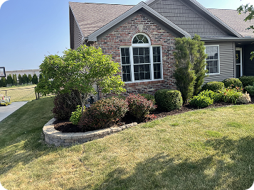 Landscaped front yard features brick home, lush shrubs, and small trees in a raised stone-edged bed under bright sunlight.