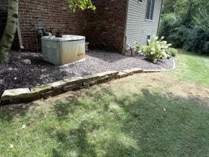 Generator sits on gravel bed beside brick house, bordered by stacked stone edging and mulch, with shrubs and shaded lawn.