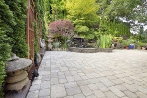 Japanese-style garden patio with stone pavers, small waterfall, bamboo fencing, statues, and lush ornamental foliage.