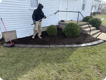 Worker spreads mulch around front bushes near concrete steps of a house, using a metal rake in a neatly edged garden bed.