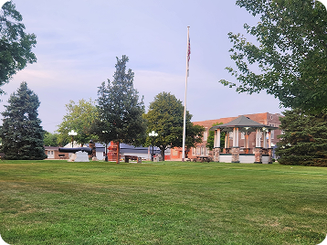Town square lawn with gazebo, flagpole, trees, benches, and historical cannons, surrounded by buildings under a soft evening sky.