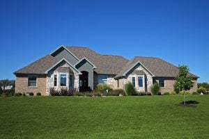 Single-story ranch-style house with brick and stone facade, large manicured lawn, and bright blue sky in the background.