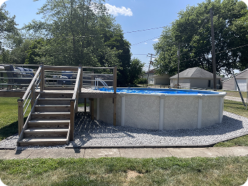 Above-ground pool with clear water sits on gravel base, with attached wooden deck and stairs, backed by trees and utility lines.