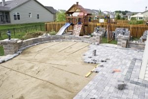 Backyard patio under construction with sand base, stacked pavers, brick retaining wall, and children\'s playset in background.