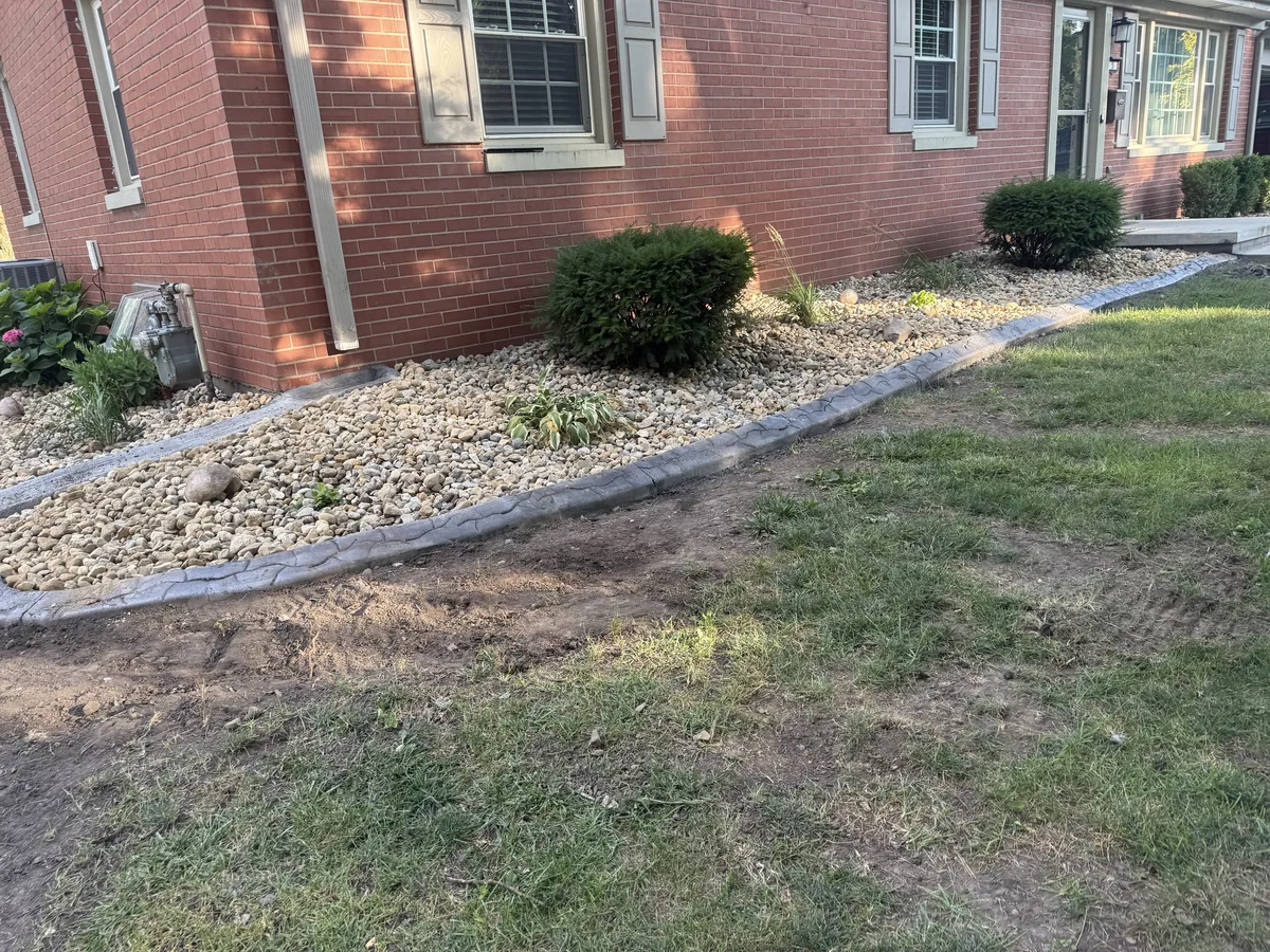 Light-colored gravel and a concrete drainage trench line a red brick wall, with scattered plants and downspout extension.