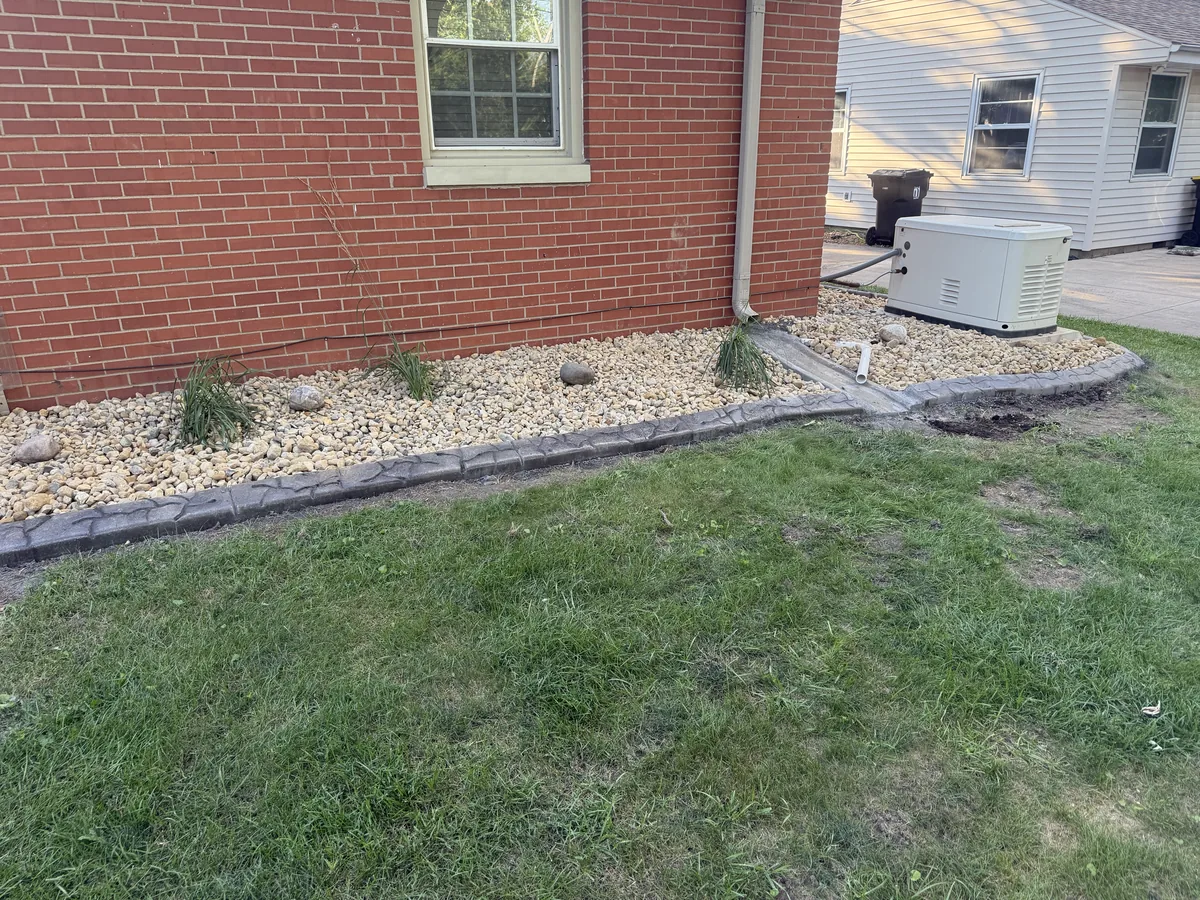 Stone-edged garden bed surrounds a home generator, with dark gravel mulch, brick siding, and flowering hydrangeas.