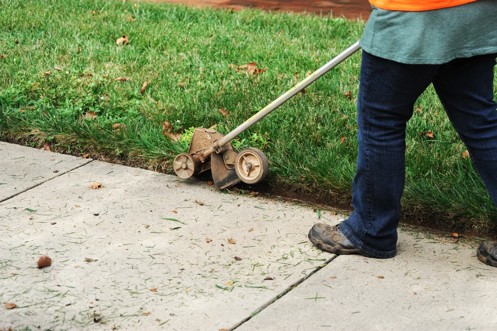 Worker edges grass along a sidewalk showing where landscape curbing can outlast traditional edging.
