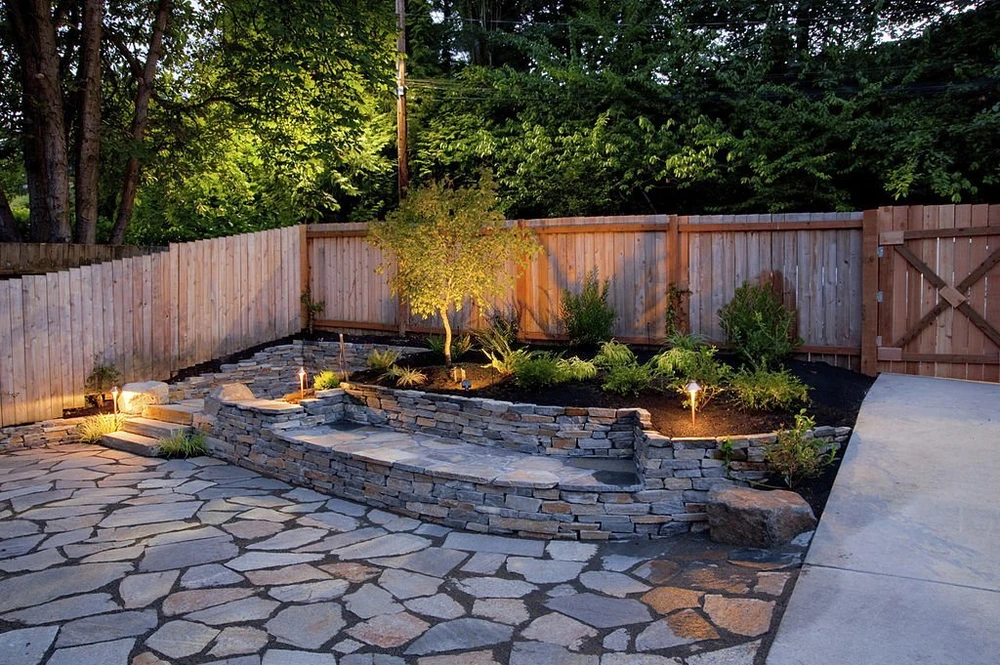 Warm garden lights highlight a stone patio with raised beds and a wooden privacy fence.