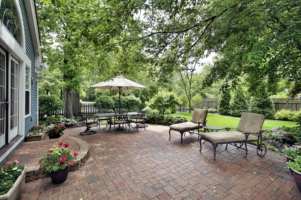 Brick patio with lounge chairs, dining table, umbrella, and garden beds under mature trees for classic hardscaping.