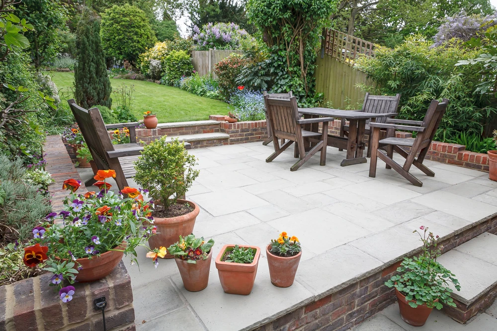 Spacious gray paver patio with a wooden dining set, terracotta flowerpots, brick-edged beds, lush shrubs, tall trees, and a slatted garden fence.