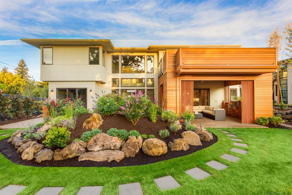 Landscaping surrounds a modern wood and white home with boulders and purple flowers beside curved stone steps.