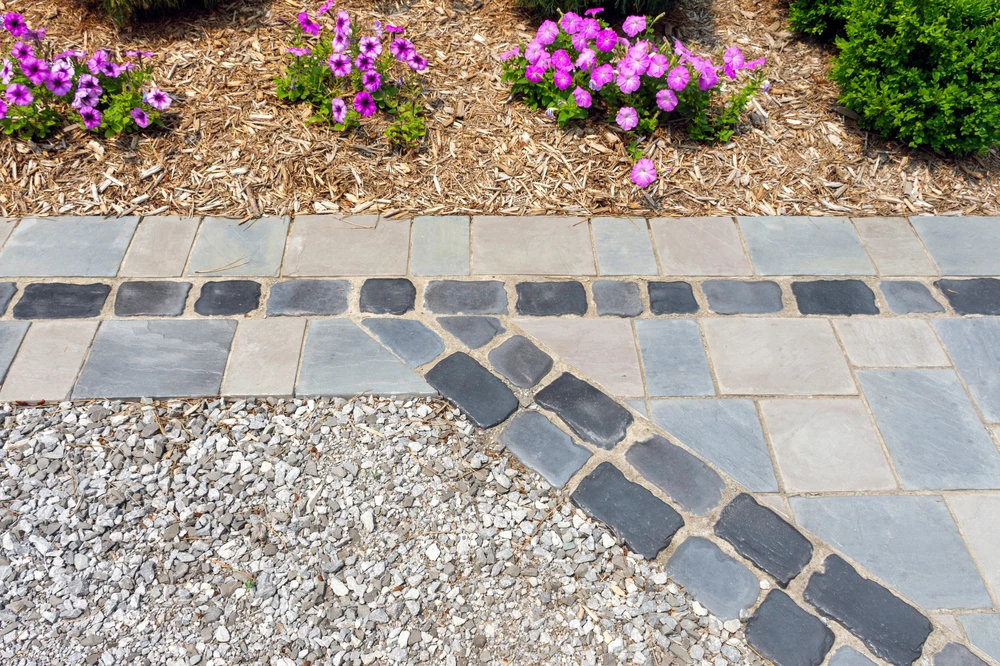 Detailed hardscaping blends pale slate and dark cobblestones beside purple flowers and tan mulch.