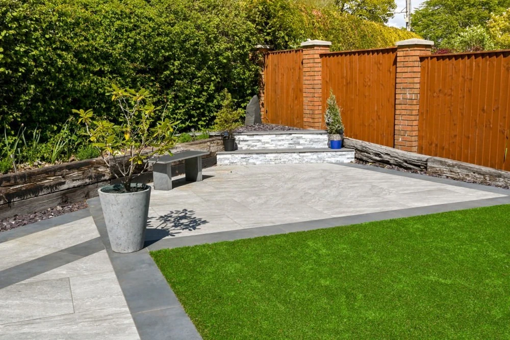Sleek gray hardscaping frames a raised patio with steps, planters, a wood fence, and vivid green lawn.