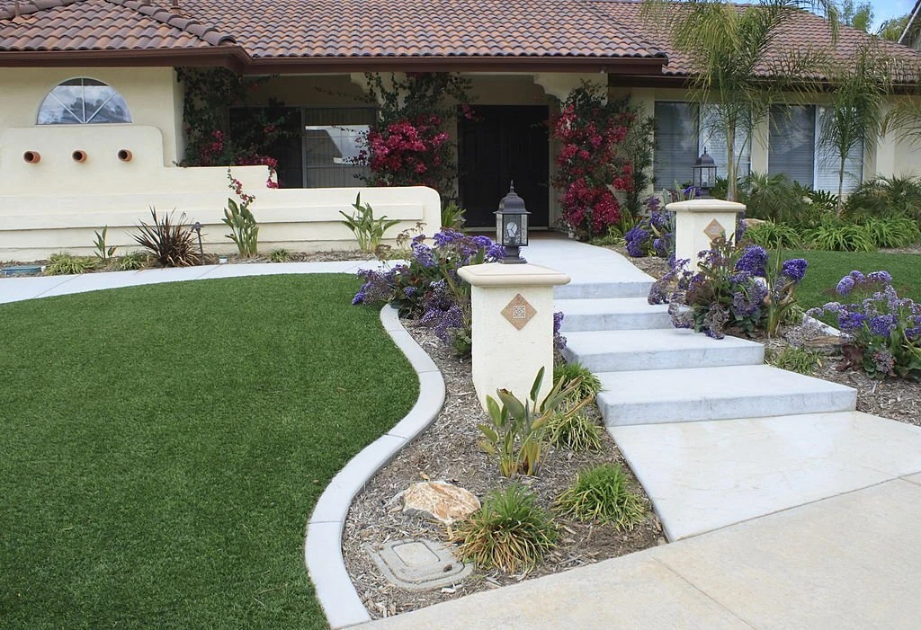  White stucco home entrance with broad concrete steps, a curved green lawn, purple flower beds, palms, flowering vines, and lantern-topped pillars.