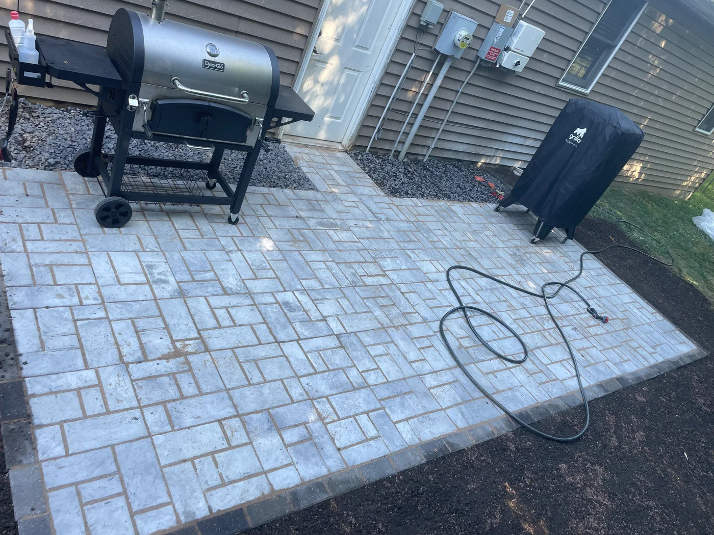 Paver patio with a grill beside a vinyl-sided home and gravel landscape beds.