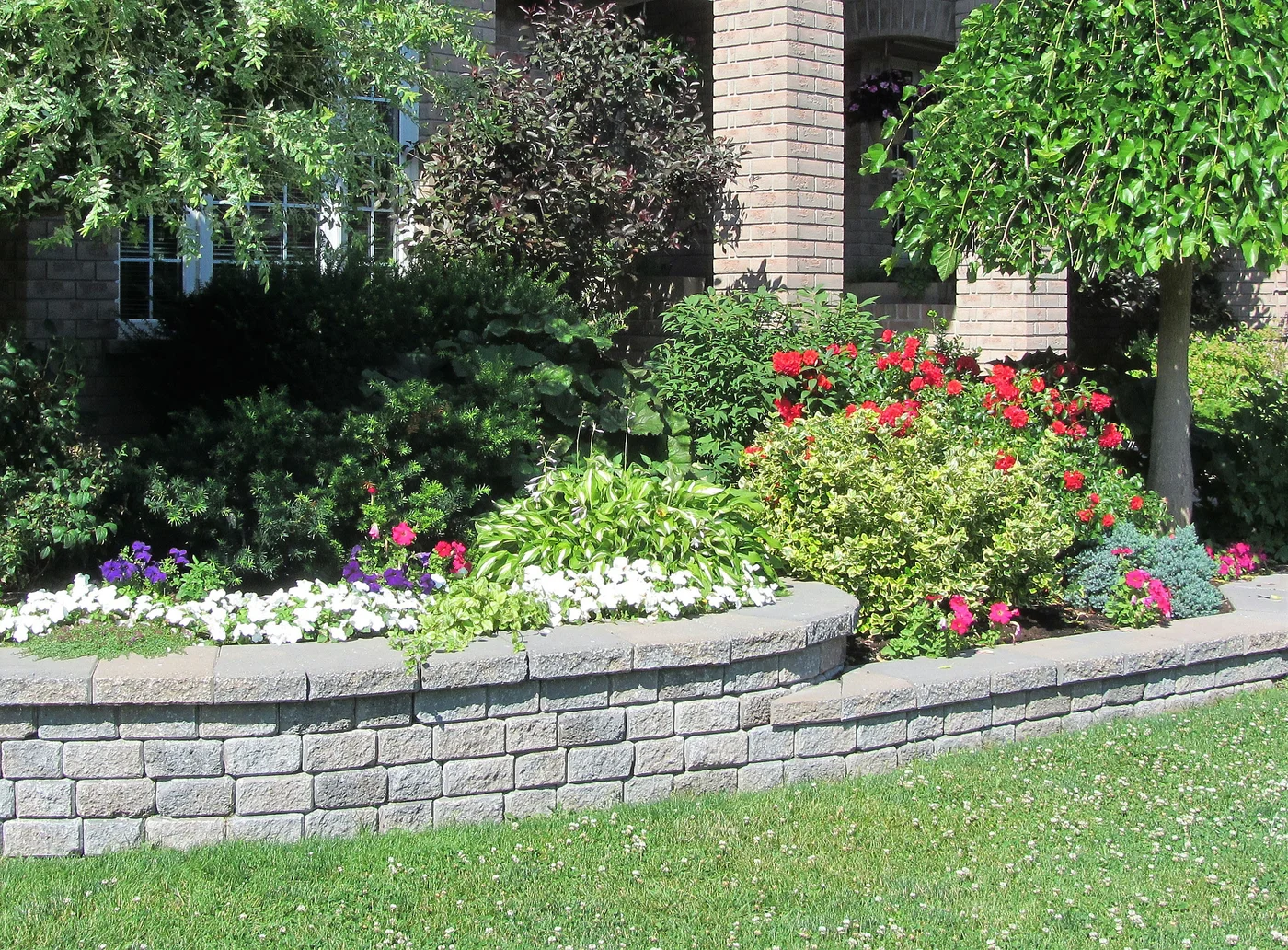 Curved gray block retaining wall borders a raised flower bed with red, white, and purple blooms beside green lawn.