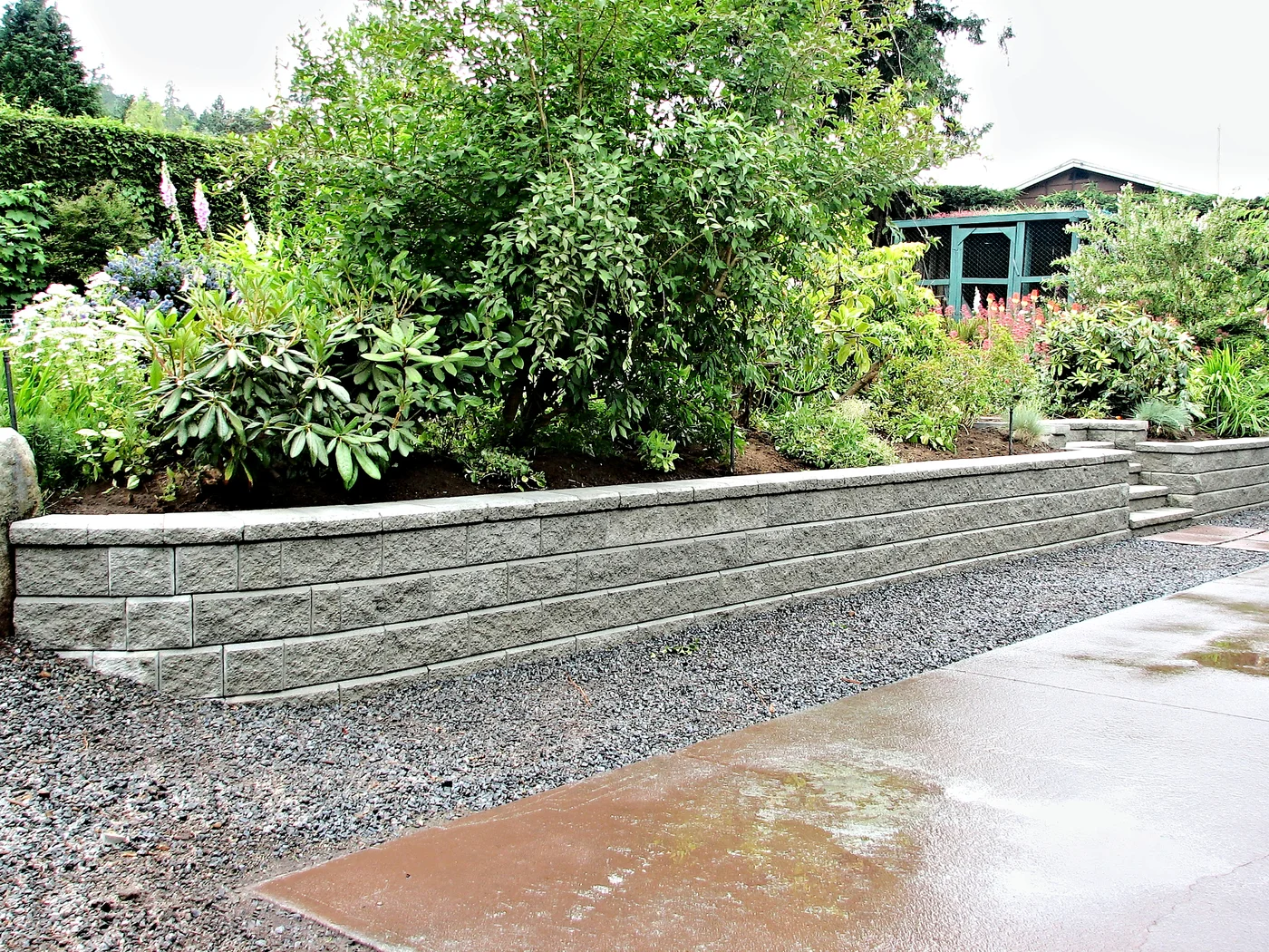 Gray block retaining wall below a densely planted slope with a gravel strip and wet concrete surface in front.