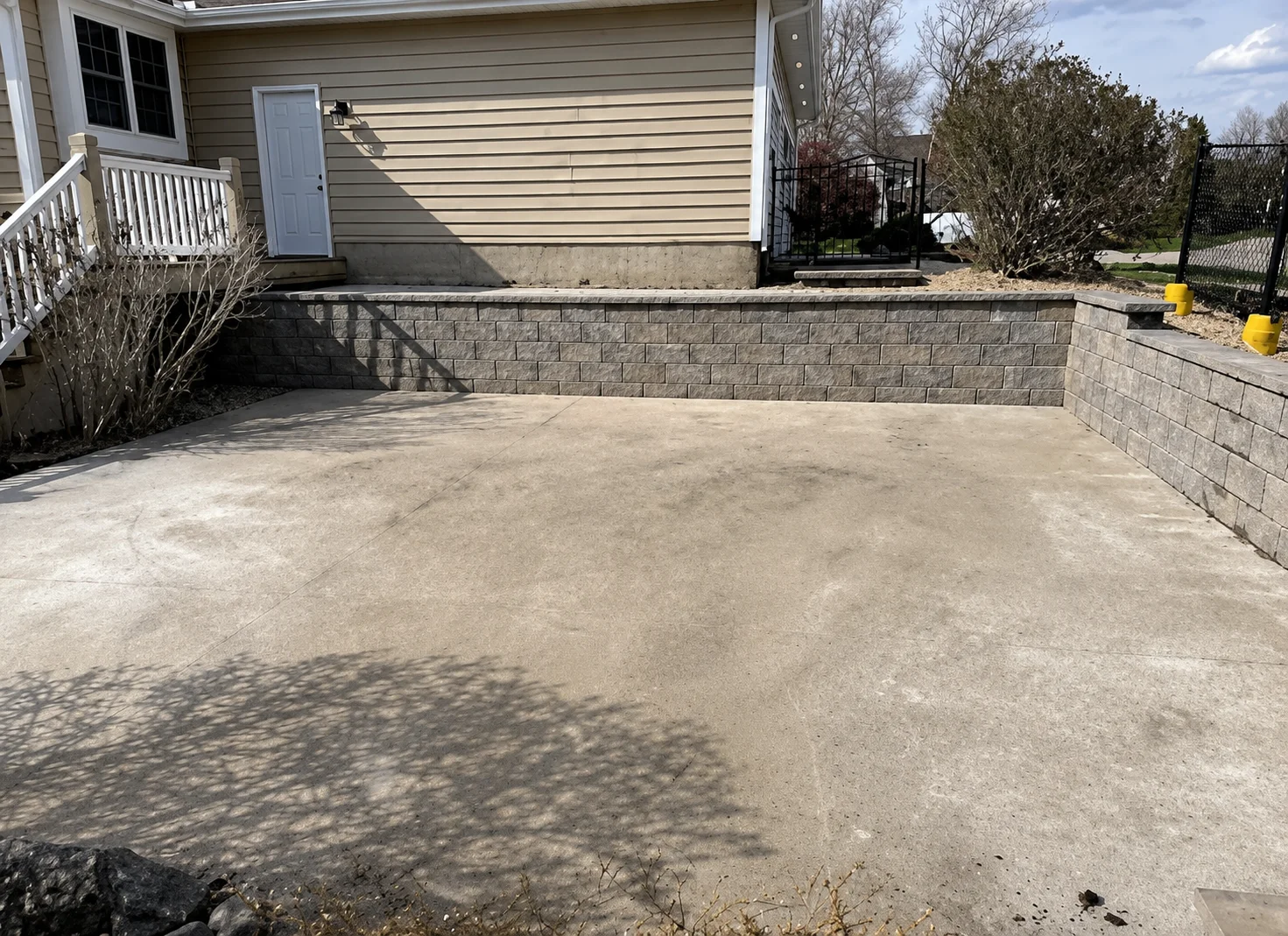 Straight gray block retaining wall borders a broad concrete patio beside a beige-sided home and black fence.