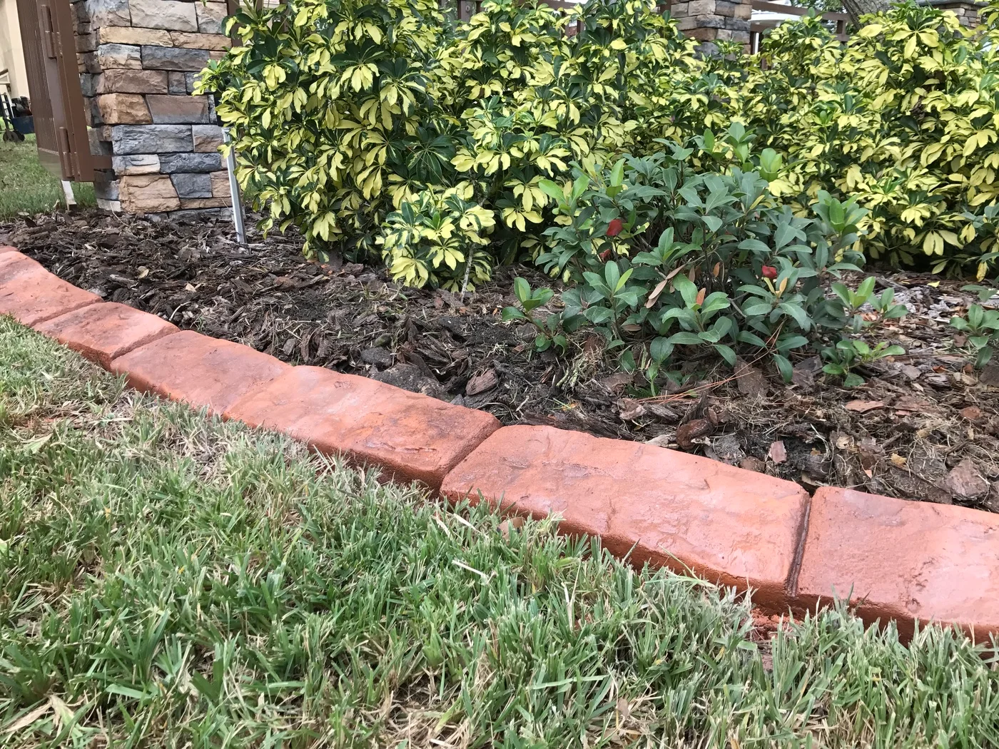 Stamped red concrete curbing beside a dark mulch landscaping bed and variegated bushes near a stone pillar.