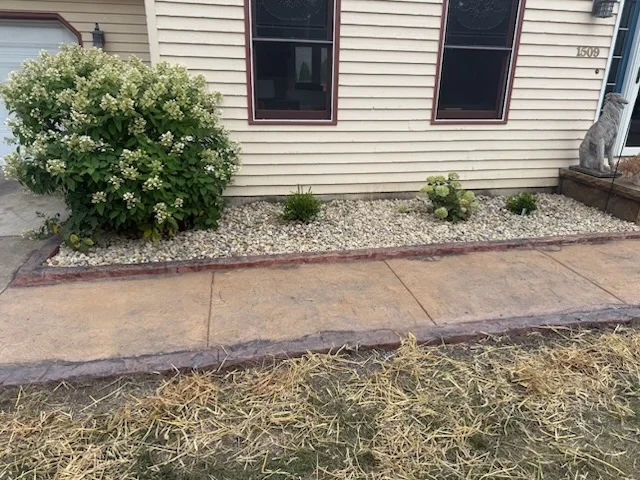 Stamped concrete walkway beside a gravel landscaping bed and a large flowering bush in front of a house.