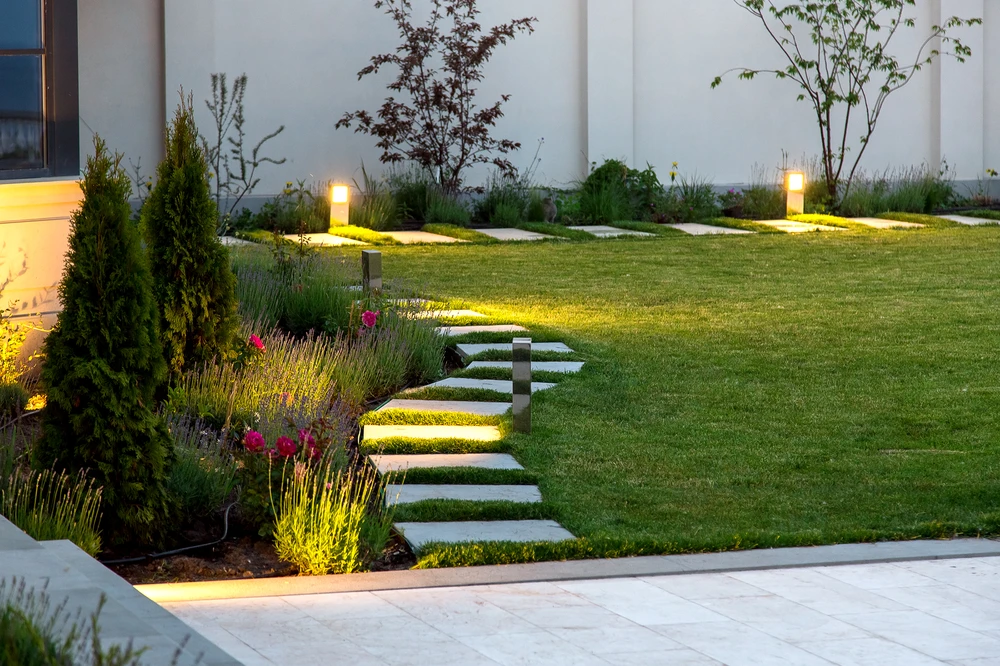 Warm landscape lighting highlights stepping stones through a green lawn beside lavender beds and young trees.