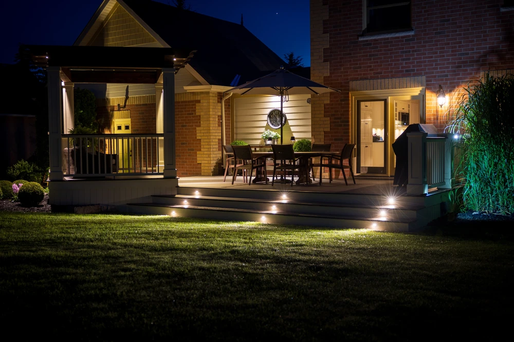 Landscape lighting outlines dark deck steps and a covered patio with warm beams against brick walls and deep blue sky.