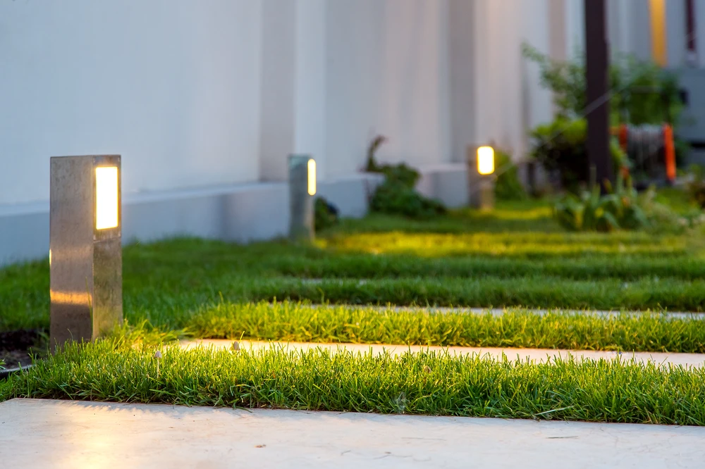 Modern landscape lighting glows from square metal posts along pale stepping stones set between strips of vivid grass.