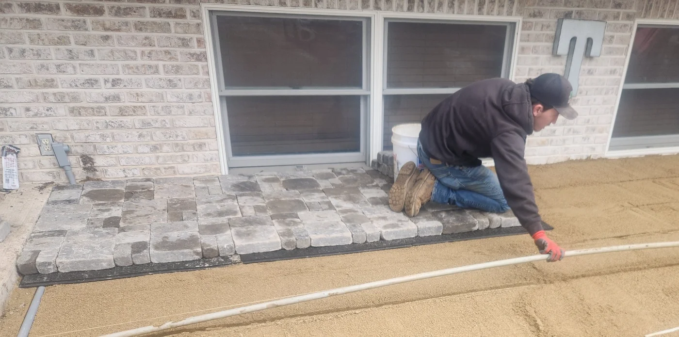 Crew member installs gray pavers beside a brick home over a prepared sand base.
