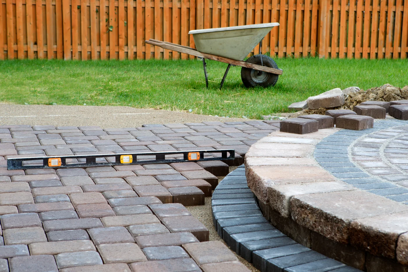 Partially installed paver patio on a sand base with a leveling tool, beside a raised circular stone feature and a wheelbarrow on the lawn.