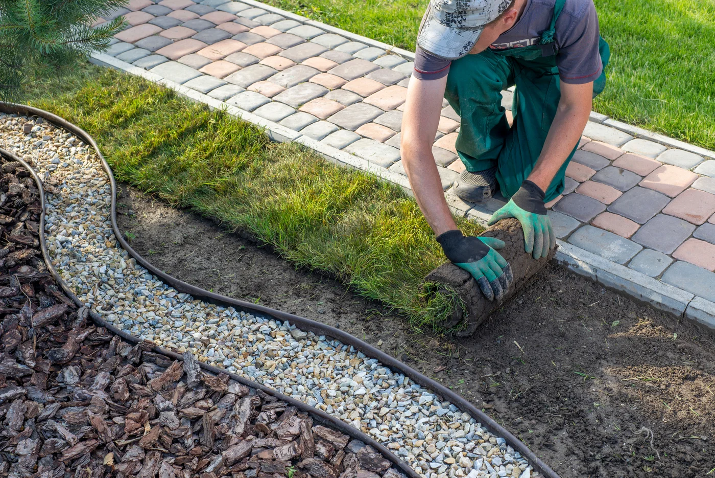 Landscaper unrolls fresh sod beside a paver walkway, gravel border, and mulched planting bed.