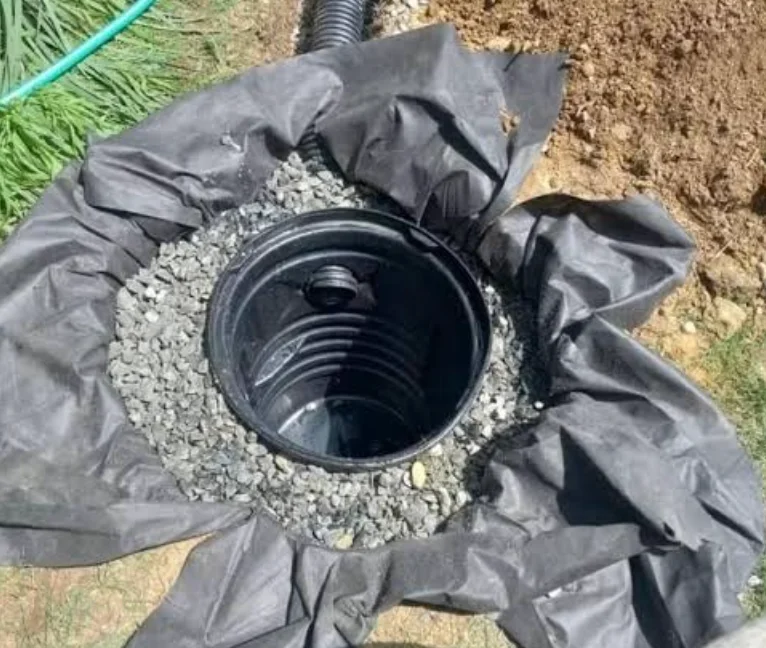 A round black catch basin sits in gray gravel inside folded black fabric beside brown soil. A corrugated pipe enters from the top.