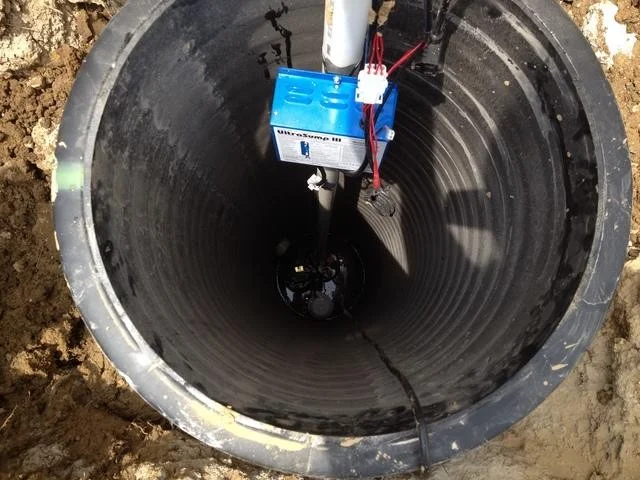 A blue pump hangs inside a deep black ribbed basin with red wires and a white pipe. Dark water and the pump sit at the bottom.